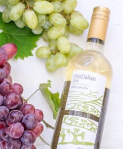 A bottle of closDalian white grenache wine lies on a white surface, surrounded by clusters of green and red grapes, a green grape leaf in the background, and a plate of stuffed dates completing the elegant scene.