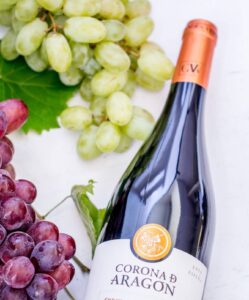 A bottle of Corona de Aragón red wine lies on a white surface next to clusters of green and red grapes with green leaves, inviting pairing with stuffed dates for a perfect appetizer.
