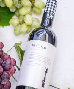 A bottle of El Circo red wine lies on a white surface next to clusters of green and red grapes, with stuffed dates and a grape leaf partially visible in the background.