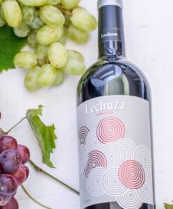 A bottle of Lechuza Garnacha wine lies on a white surface next to a bunch of green grapes, red grapes, and a plate of stuffed dates. The wine label features striking red and white concentric circles.