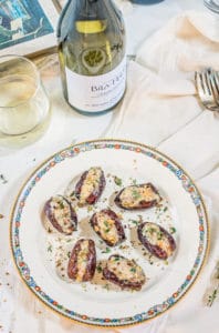 A decorative plate with stuffed dates, garnished with herbs, sits on a white tablecloth. Nearby are a glass and a bottle of white wine, two forks, and a napkin.