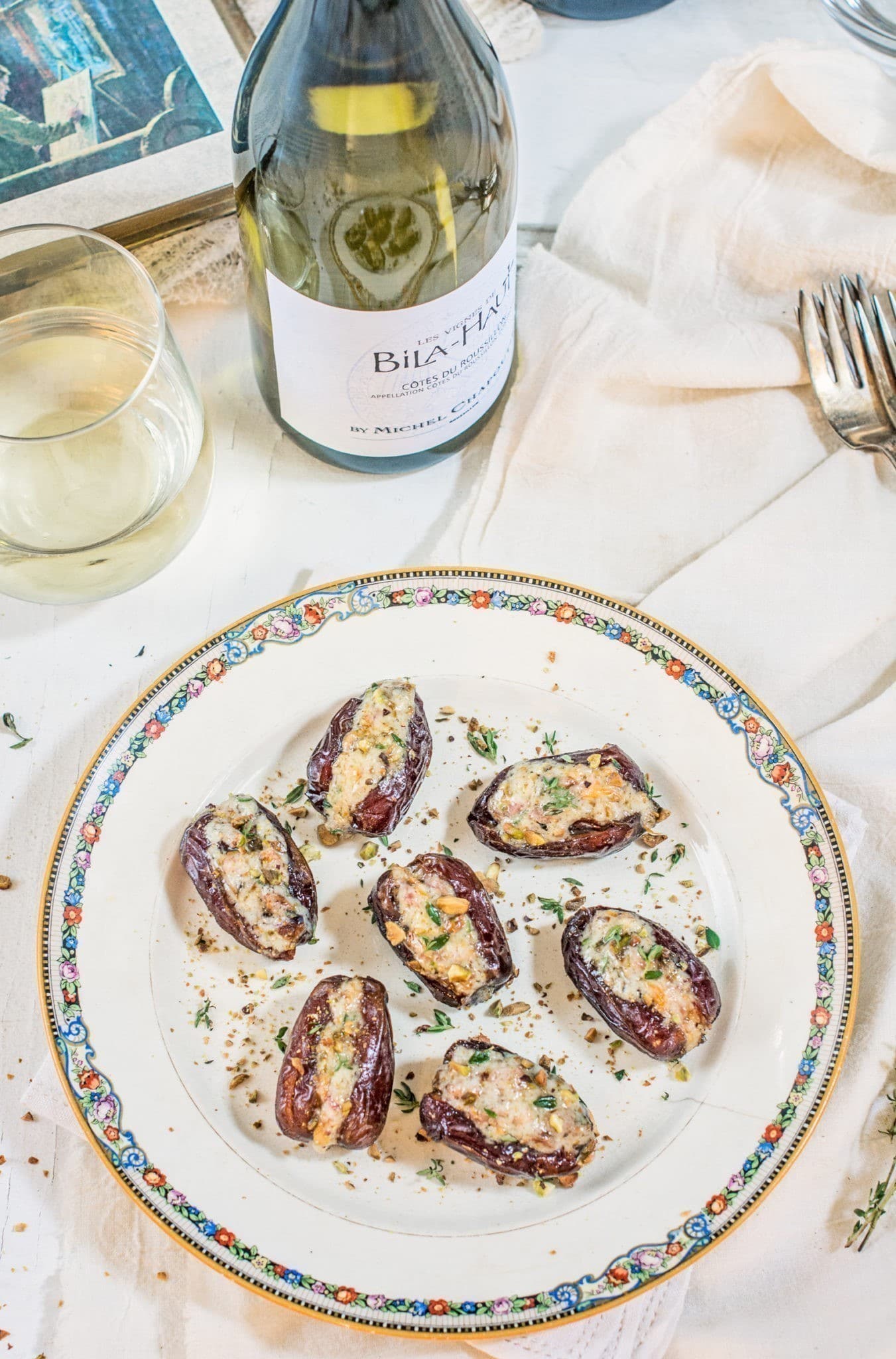 A decorative plate with stuffed dates, garnished with herbs, sits on a white tablecloth. Nearby are a glass and a bottle of white wine, two forks, and a napkin.