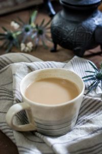 A ceramic cup filled with milky Whiskey Chai rests on a striped cloth, with thistle flowers and a dark teapot in the background.