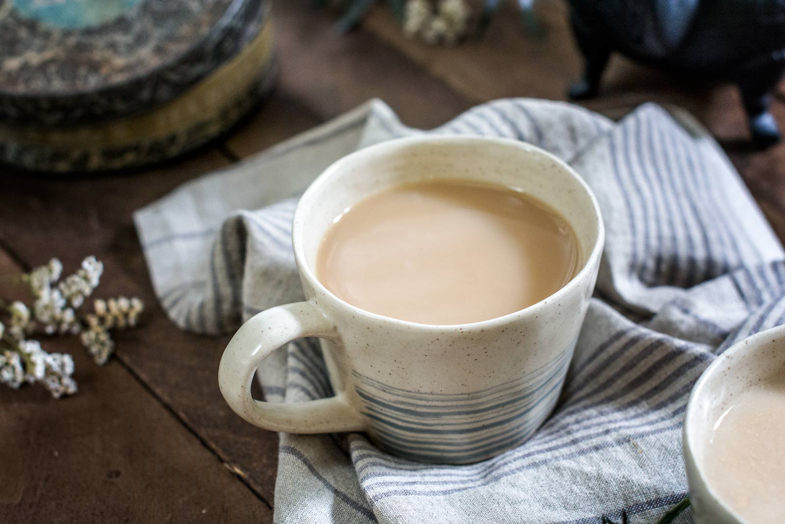 A ceramic mug filled with light brown Whiskey Chai sits on a striped cloth on a wooden table, with another partially visible cup and some decorative items in the background.