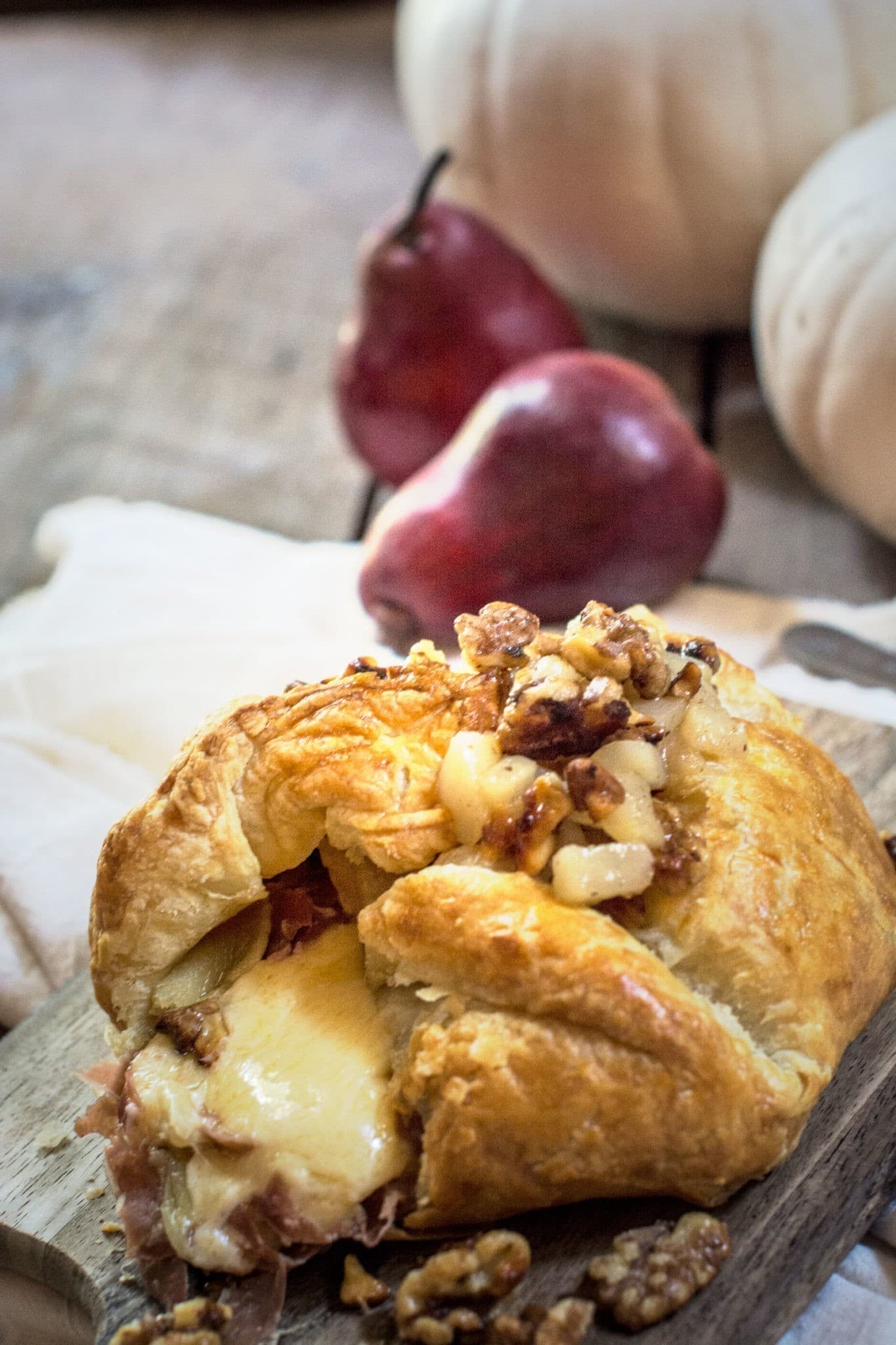 A golden, flaky Baked Gouda pastry stuffed with melted cheese, prosciutto, and walnuts sits on a wooden board. In the background, two red pears and white pumpkins are softly blurred.
