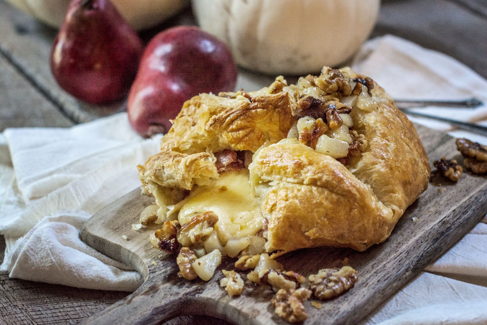 A baked Gouda puff pastry filled with melted cheese, chopped nuts, and pear pieces sits on a wooden board. Two pears and white pumpkins are in the background, with a white cloth draped nearby.