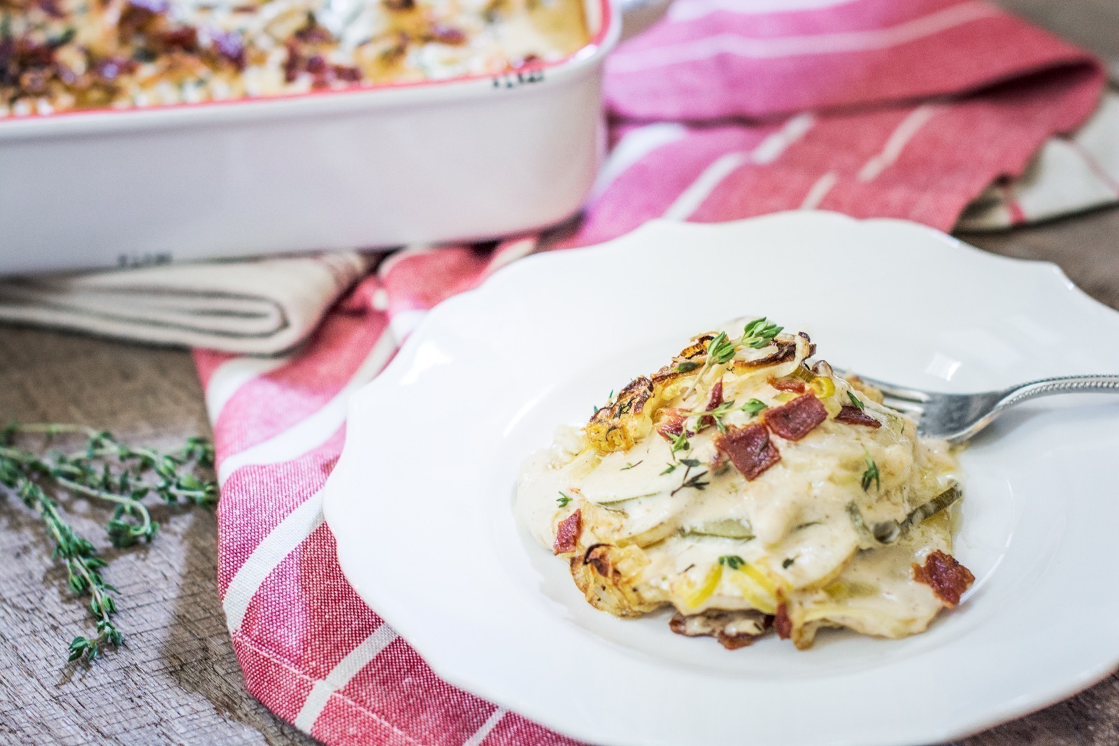A serving of creamy Zucchini Potato Gratin with herbs and crispy bacon sits on a white plate, with a fork beside it. The plate is on a red striped cloth, and the casserole dish is partially visible in the background.