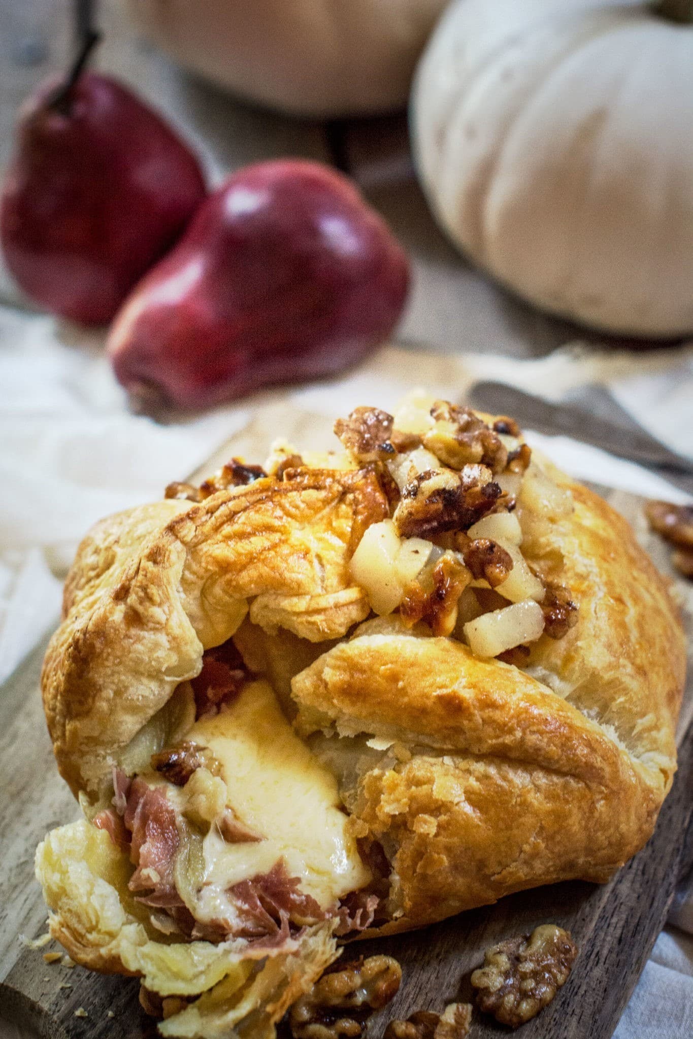 A flaky baked Gouda pastry filled with melted cheese and prosciutto, topped with chopped walnuts and pears, sits on a wooden board. In the background, two red pears and a white pumpkin are slightly out of focus.