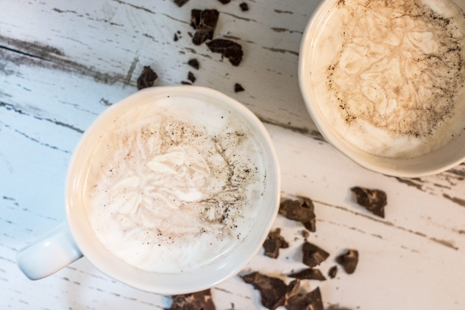 Two white mugs filled with frothy cappuccino and a hint of mocha, topped with foam and cinnamon, placed on a rustic white wooden surface with scattered pieces of broken chocolate nearby.