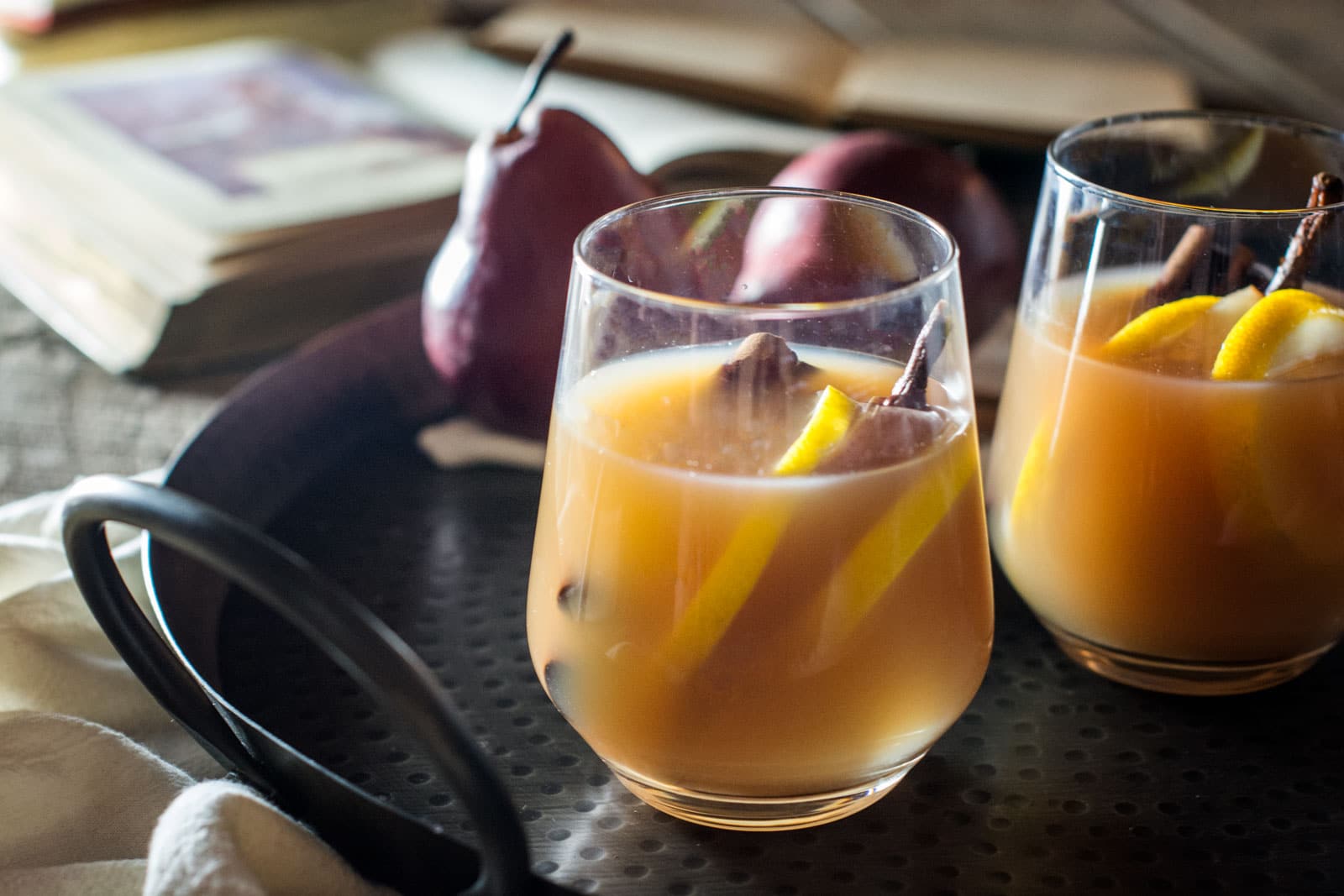 Two glasses of spiced pear cocktail, reminiscent of pear cider, with lemon slices and cinnamon sticks on a dark tray, surrounded by whole pears and an open book in the background.