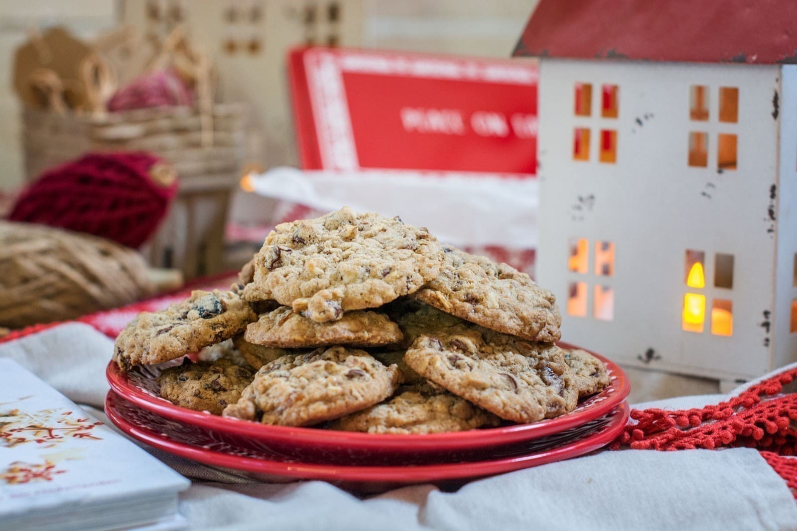 A plate of Everything Cookies sits on a red dish, surrounded by festive holiday decorations, including a small white house lantern with a glowing light and a red crocheted mat.