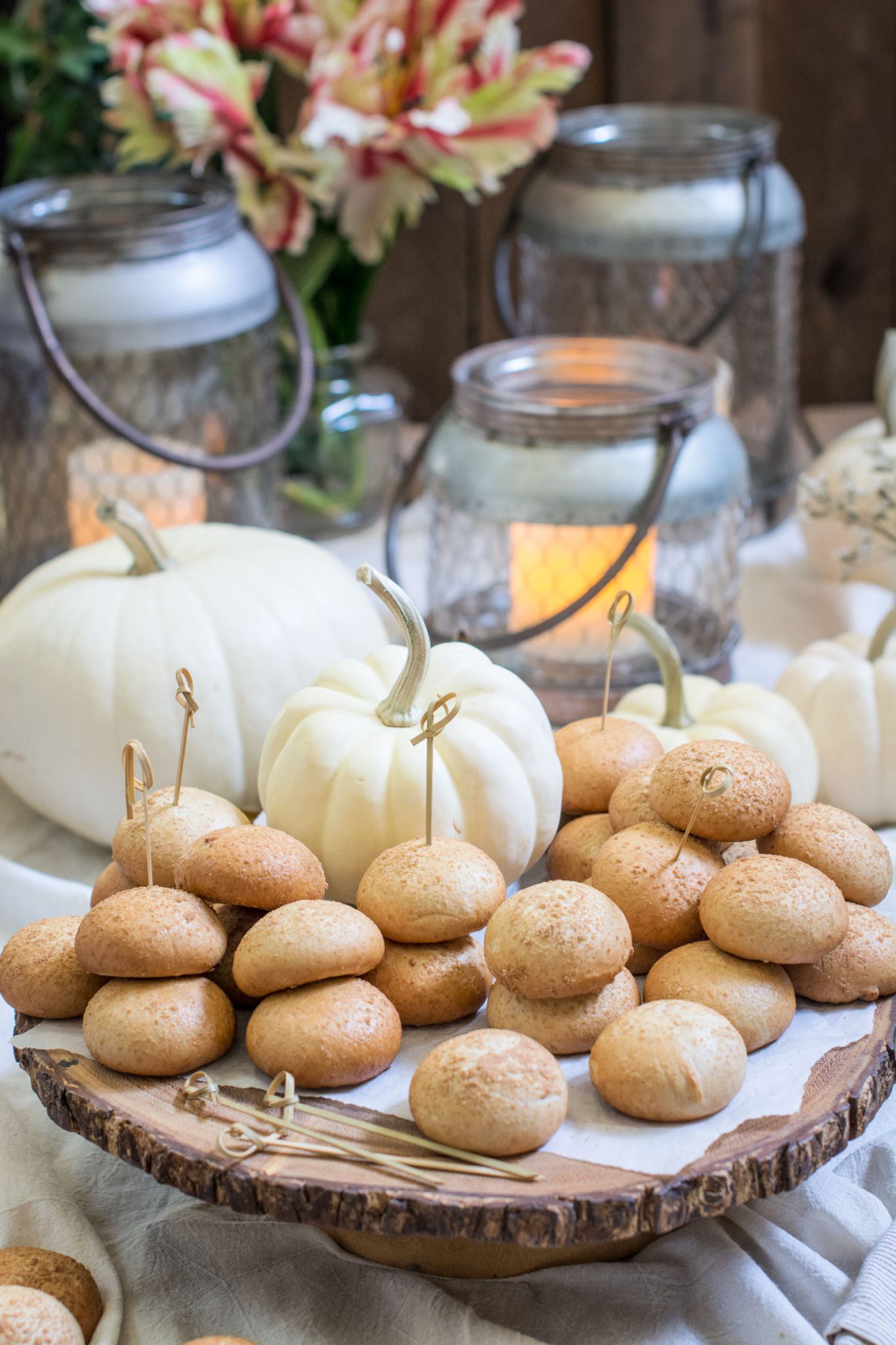 A rustic wooden tray displays stacks of round cookies with toothpicks, surrounded by white pumpkins, glass lanterns with lit candles, and flowers in the background, perfect for a cozy autumnal Holiday Brunch setting.