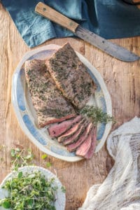 Sliced roast beef with an herb crust rests on an oval platter, garnished with fresh thyme, atop a wooden table beside a knife, cloth, and a small bowl of microgreens.