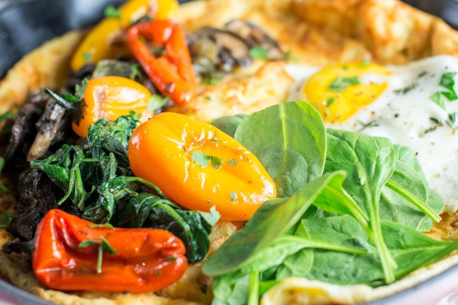 A close-up of a Savory Dutch Baby omelette topped with sautéed mushrooms, cherry tomatoes, spinach, and fresh herbs. The colorful vegetables add vibrancy and freshness to the dish.