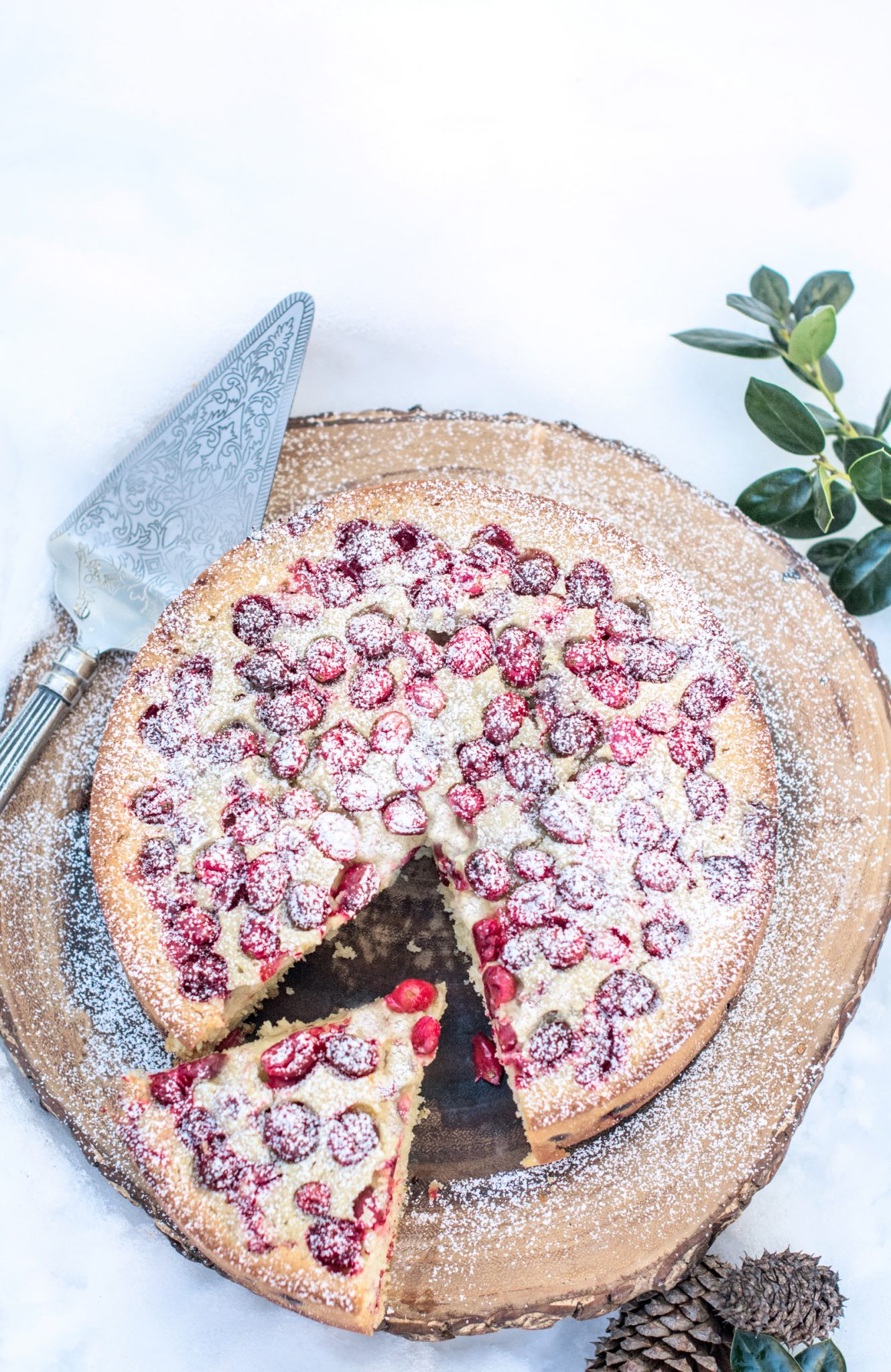 A round Olive Oil Cake dusted with powdered sugar sits on a wooden board, with one slice pulled out. A pie server rests nearby, and green leaves and pinecones decorate the snowy background.