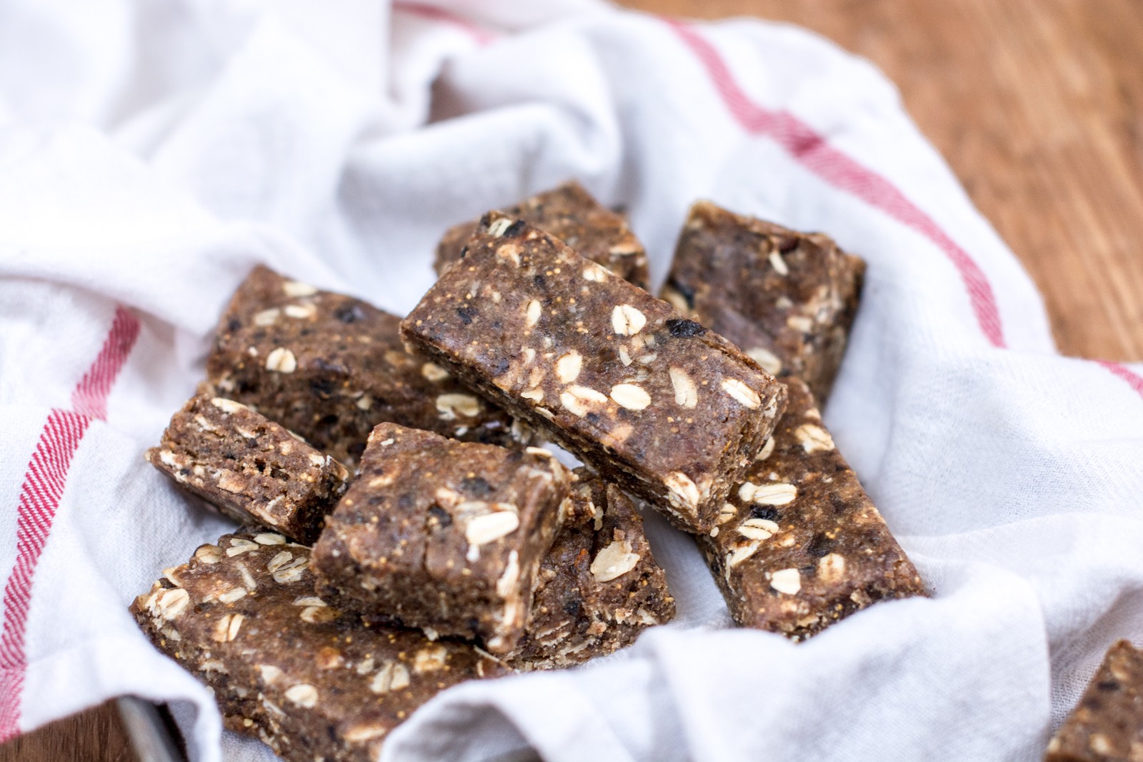 Homemade fig bars with visible oats and seeds are stacked on a white cloth with a red stripe, set on a wooden surface.