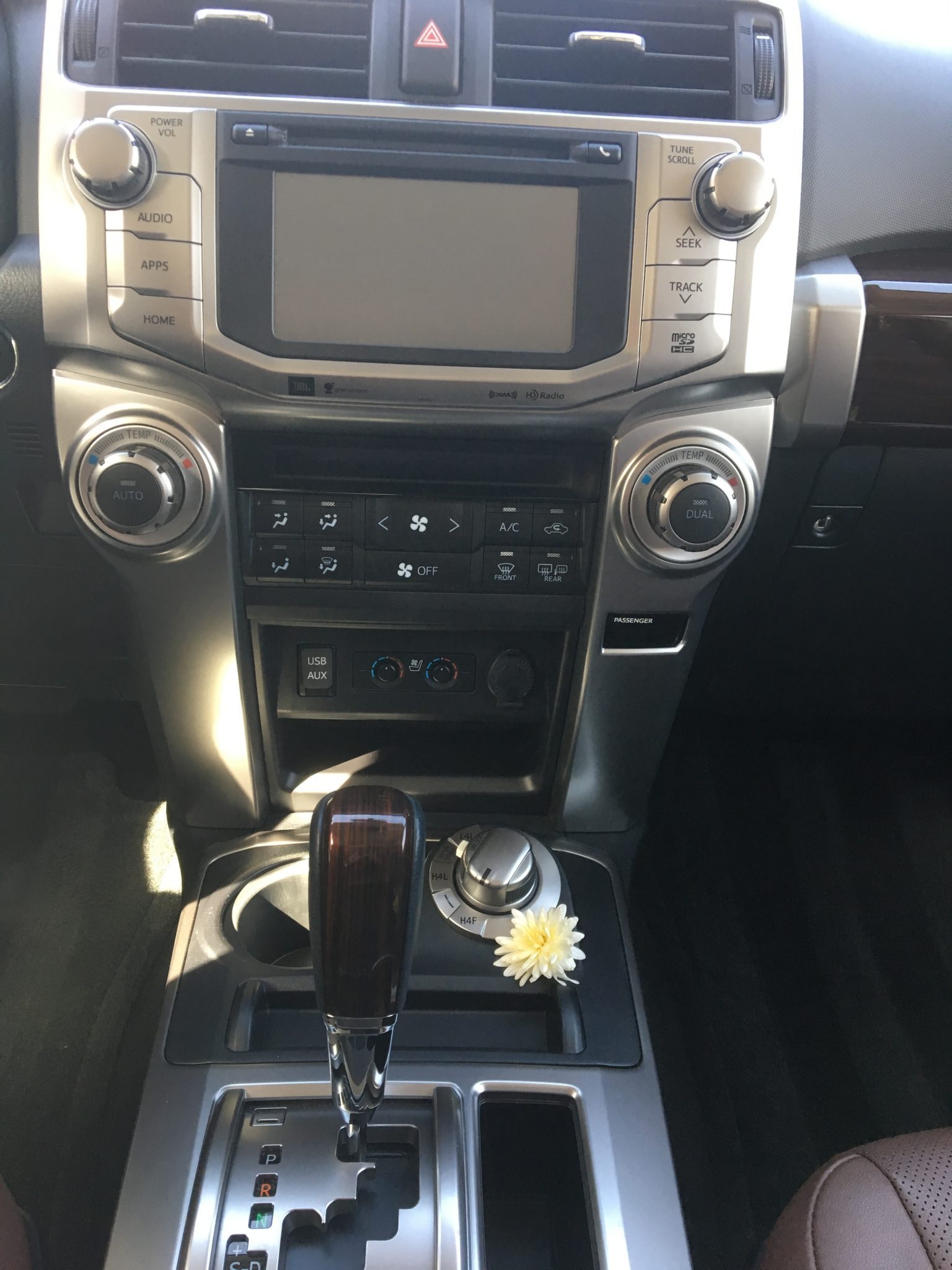 The center console of a car with a touchscreen, control buttons, gear shift, cup holders with fig bars tucked inside, and a small white artificial flower beside the gear lever. Sunlight highlights parts of the interior.