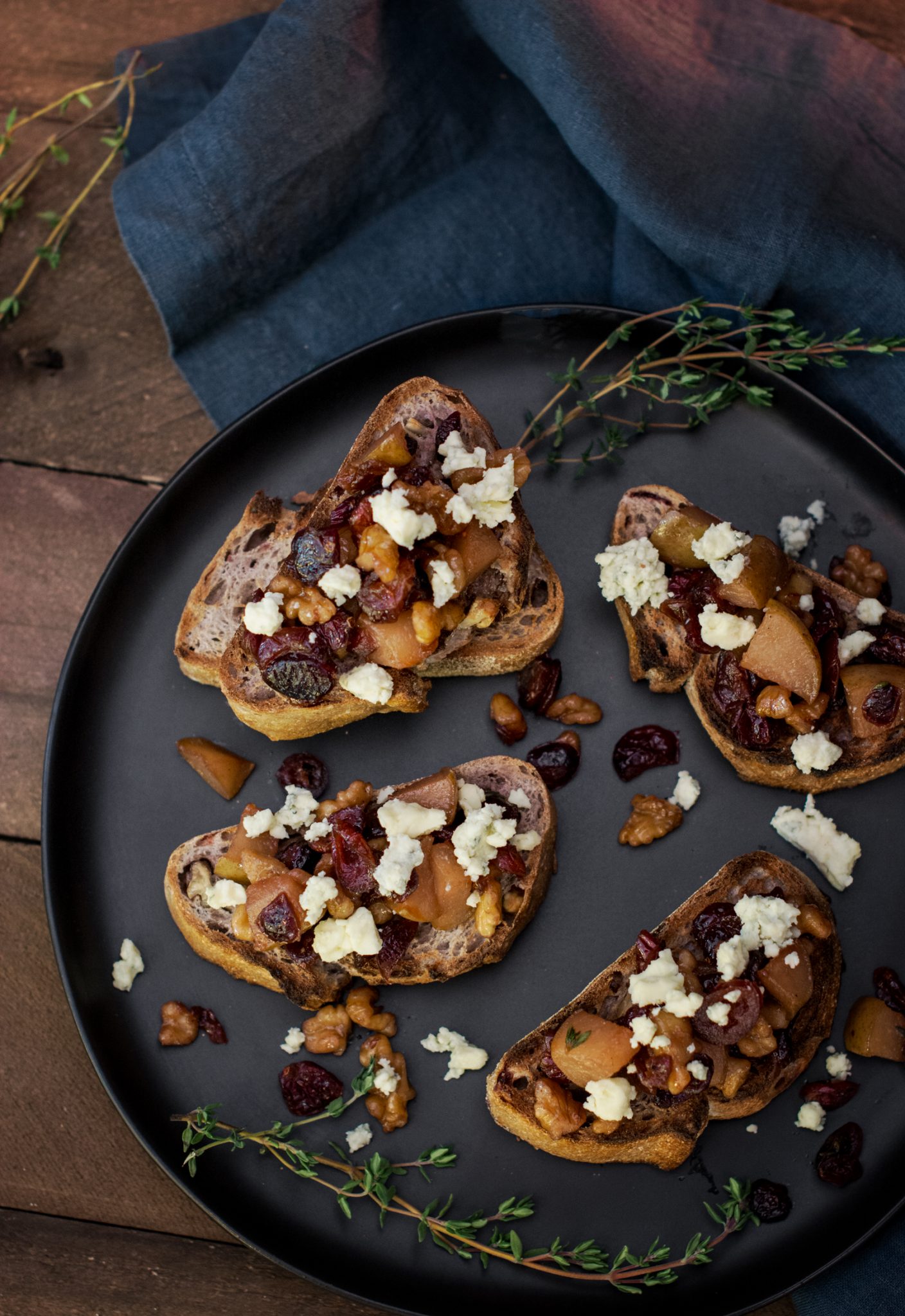 A black plate holds Blue Cheese Bruschetta—slices of toasted bread topped with crumbled cheese, chopped walnuts, dried cranberries, and caramelized pears, garnished with fresh thyme. A dark blue cloth is in the background.