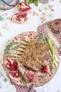 A wooden board displays a Pistachio Crusted Rack of Lamb garnished with thyme, green onions, and pomegranate seeds. Pomegranate halves and pistachios are scattered around, with a knife and a patterned napkin beside the board.