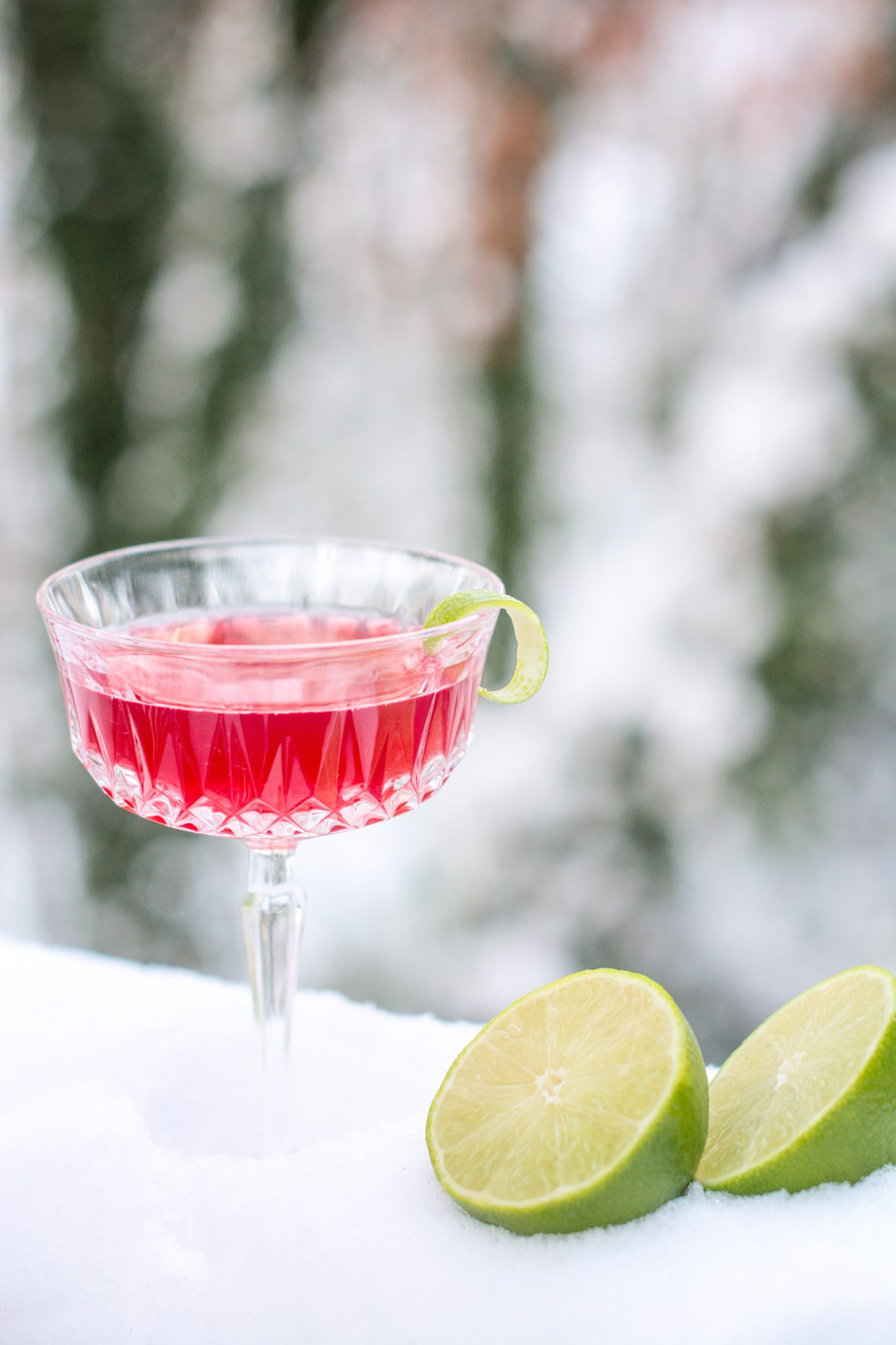 A Pomegranate Daisy pink cocktail in a crystal glass garnished with a lime twist sits on snow, with two lime halves beside it. The background is blurred with wintry trees.