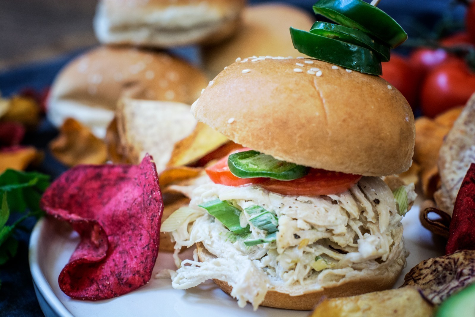 A shredded chicken slider topped with tomato and sliced jalapeños sits on a plate with colorful vegetable chips. The slider is held together with a skewer, and more sliders and tomatoes can be seen in the background.