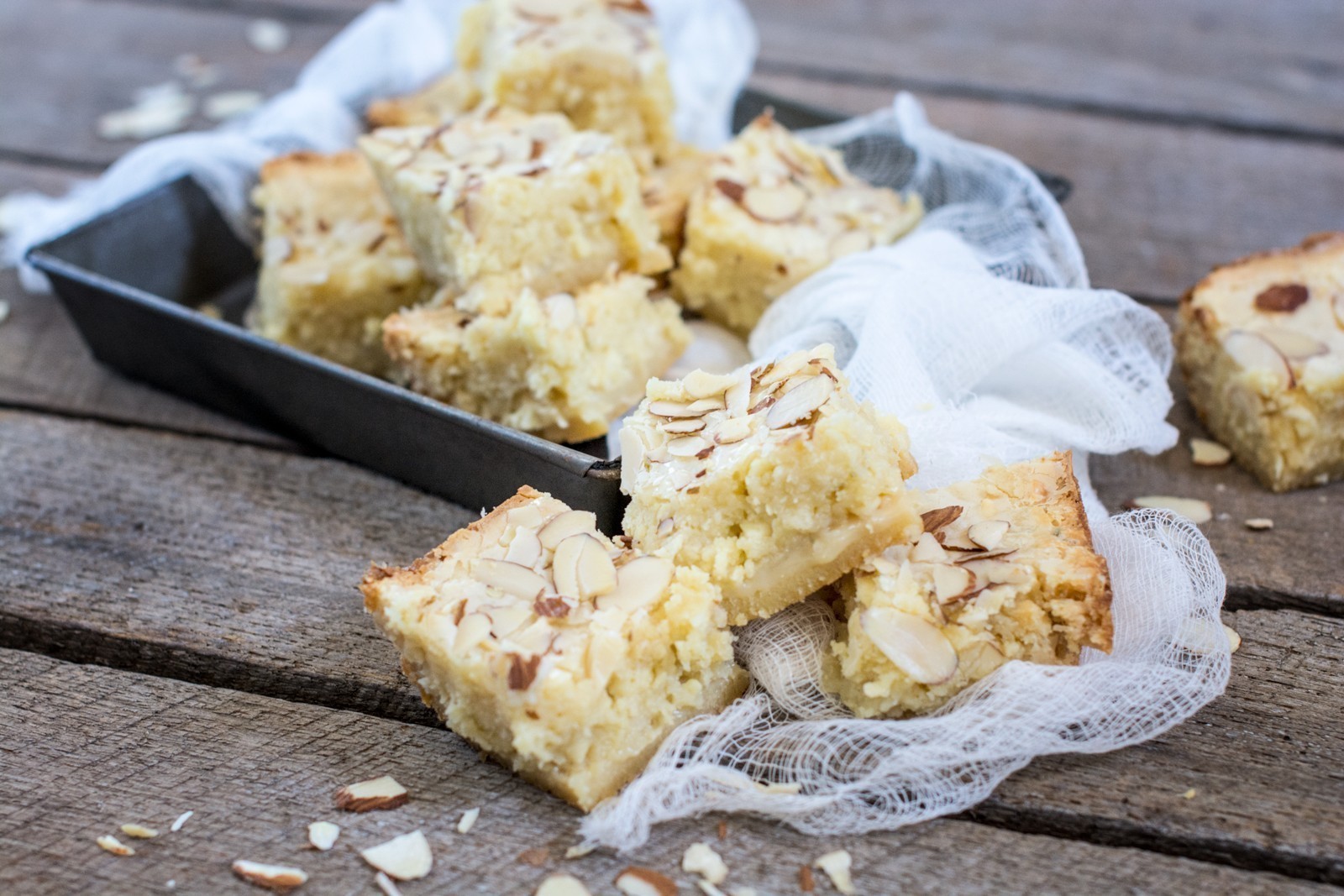 Several almond-topped blondies are arranged on a rustic wooden surface, with some in a baking tray and others stacked on white cloth. Sliced almonds are scattered around the moist, golden bars.