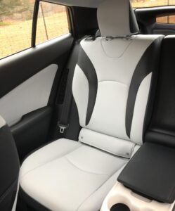 Back seat of a car with modern two-tone white and black upholstery, cup holders in the center armrest—perfect for holding drinks or blondies—and a seatbelt visible on the left. A window reveals grass and trees outside.