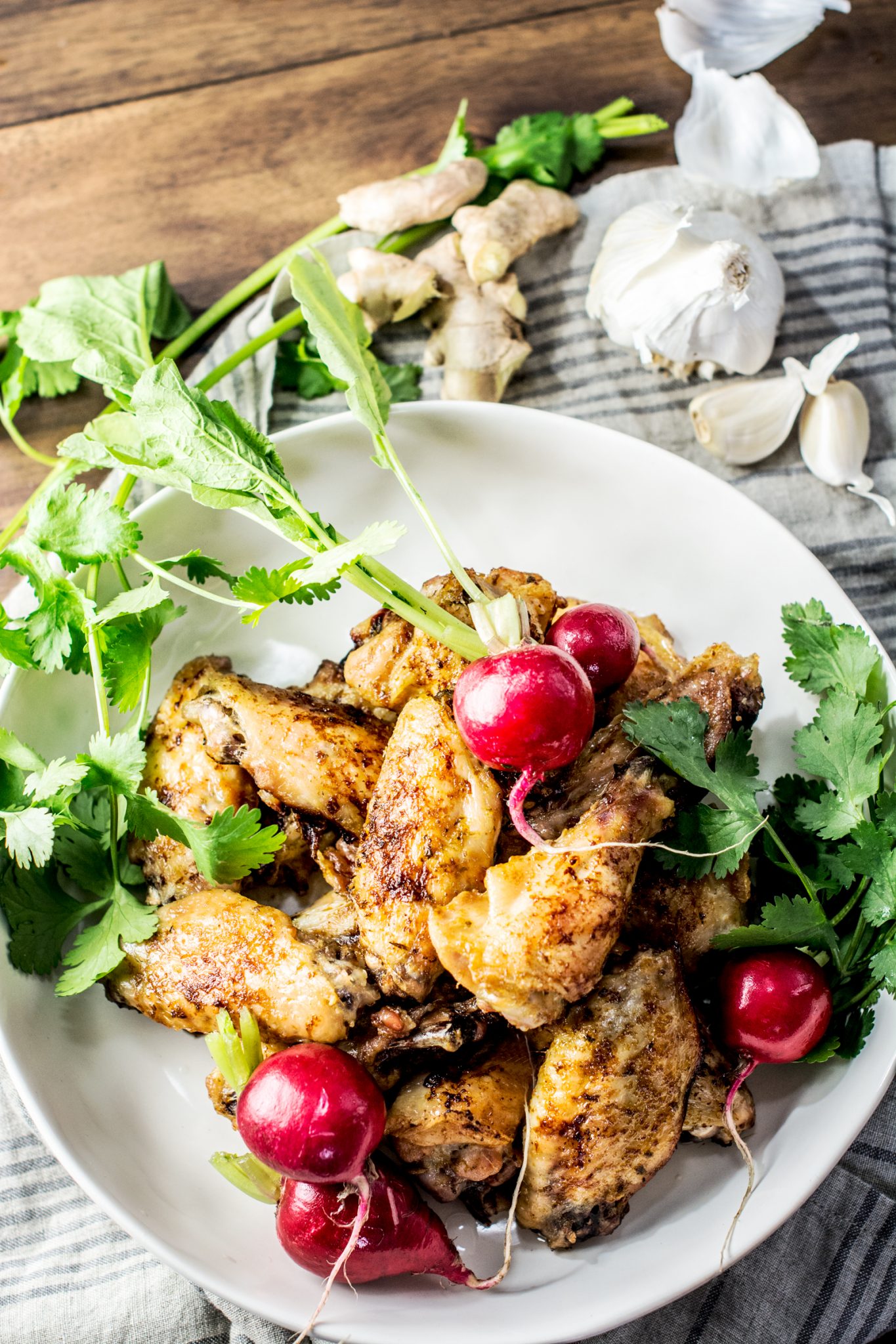 A white bowl filled with grilled wings, garnished with fresh cilantro and whole red radishes, sits on a striped cloth with garlic, ginger, and more cilantro in the background.