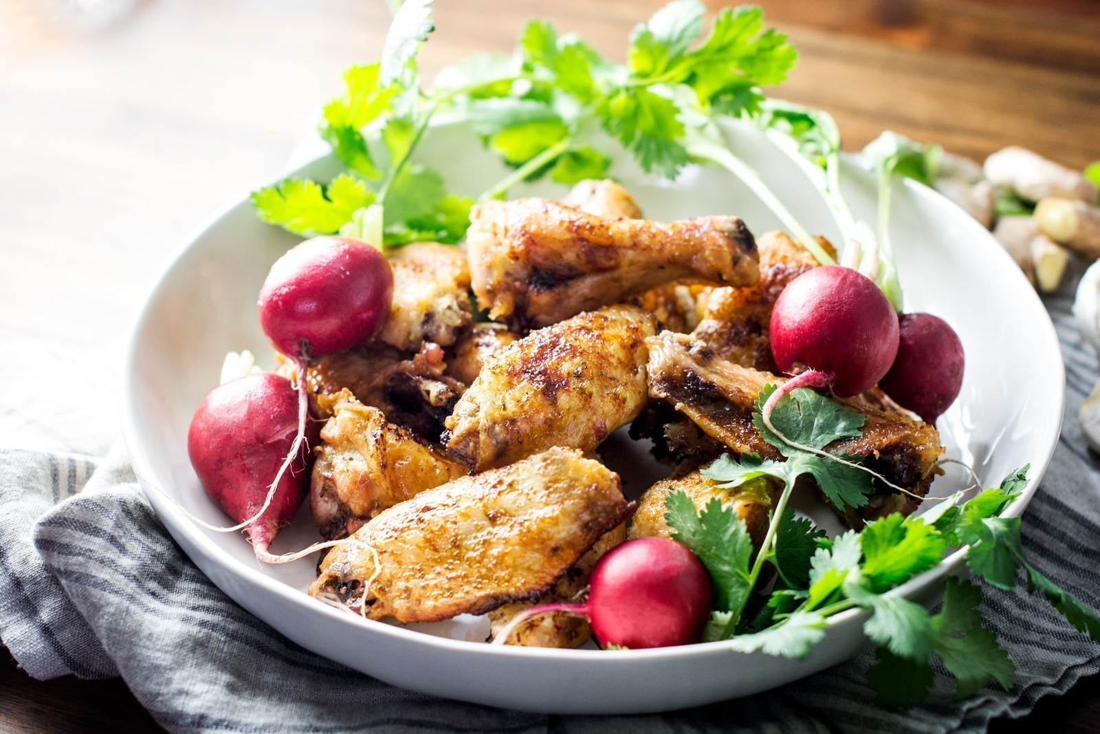 A white bowl filled with crispy wings, fresh radishes, and sprigs of cilantro, placed on a striped cloth with a wooden table in the background.
