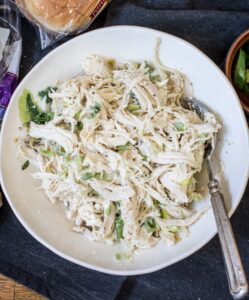 A bowl of shredded chicken salad with green peppers sits on a dark cloth, surrounded by sliders made with sesame seed buns, sliced jalapeños in a small dish, and whole jalapeños. A spoon rests in the bowl.