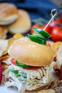 A shredded chicken slider on a sesame bun, topped with lettuce, tomato, and sliced jalapeños, secured with a toothpick. Surrounding the slider are chips and blurred tomatoes in the background.