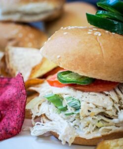 A shredded chicken slider topped with tomato and sliced jalapeños sits on a plate with colorful vegetable chips. The slider is held together with a skewer, and more sliders and tomatoes can be seen in the background.