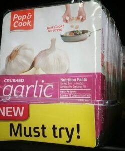 A package of Pop & Cook crushed garlic is displayed on a store shelf with a yellow sign below reading NEW Must try! The label features garlic bulbs, a pan with food—perfect for adding bold flavor to wings.