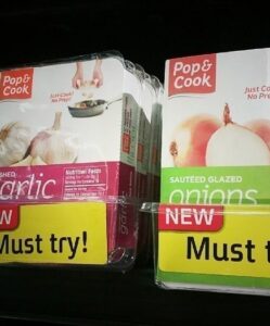 Two boxes of Pop & Cook frozen seasoning cubes, one labeled Crushed Garlic and the other Sautéed Glazed Onions, are displayed on a store shelf with NEW and Must Try! signs—perfect for adding bold flavor to your wings.