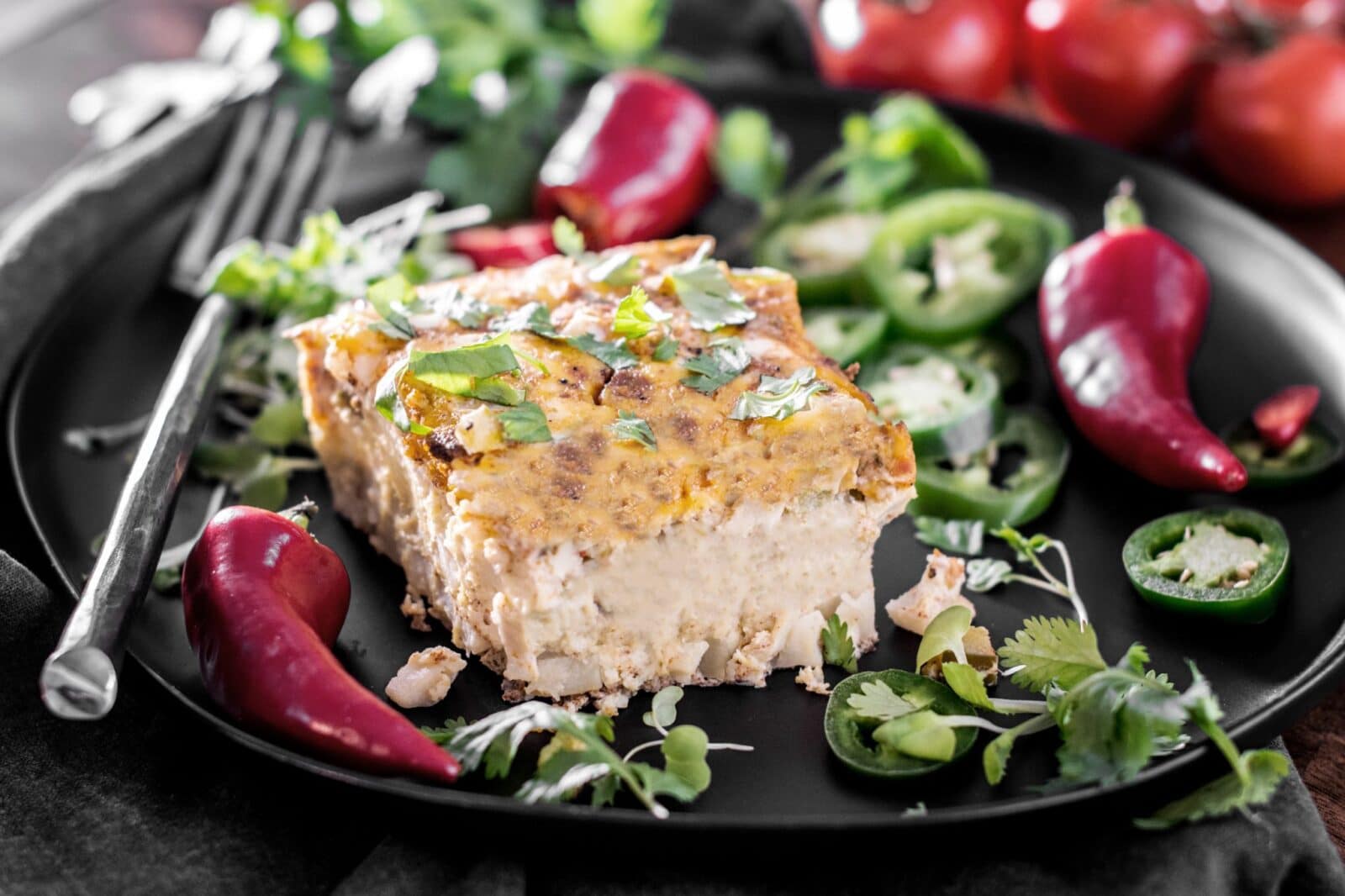 A slice of creamy casserole topped with herbs on a black plate, surrounded by sliced jalapeños, whole red chili peppers, fresh cilantro, and tomatoes in the background. A fork rests beside the casserole.