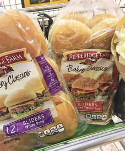 Two packs of Pepperidge Farm Bakery Classics slider buns and a bouquet of white and green flowers sit in a shopping cart. One pack holds 12 classic sliders, while the other features delicious sesame-topped buns.