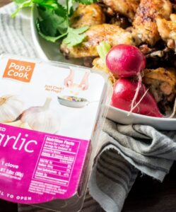 A container of Pop & Cook crushed garlic and chopped cilantro sits on a striped towel next to a plate of delicious wings garnished with herbs and radishes on a wooden table.