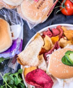 A plate with a sandwich topped with a jalapeño slice sits next to vibrant vegetable chips. Nearby are sliders buns, fresh tomatoes on the vine, and cilantro on a dark surface.