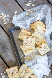 Several almond-topped blondies are arranged on a metal tray and rustic wooden surface, with white gauze fabric draped around them. Sliced almonds are scattered on and around the bars.