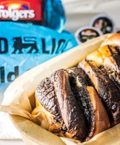 A loaf of Coffee Babka bread in parchment paper sits in front of a blue Good & Gather cold cup package and a container of Folgers coffee, with coffee pods in the background.