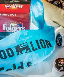 A Food Lion plastic bag covers a box of Folgers Black Silk Keurig coffee pods, with several pods scattered nearby and a coffee babka in a paper tray in the background.