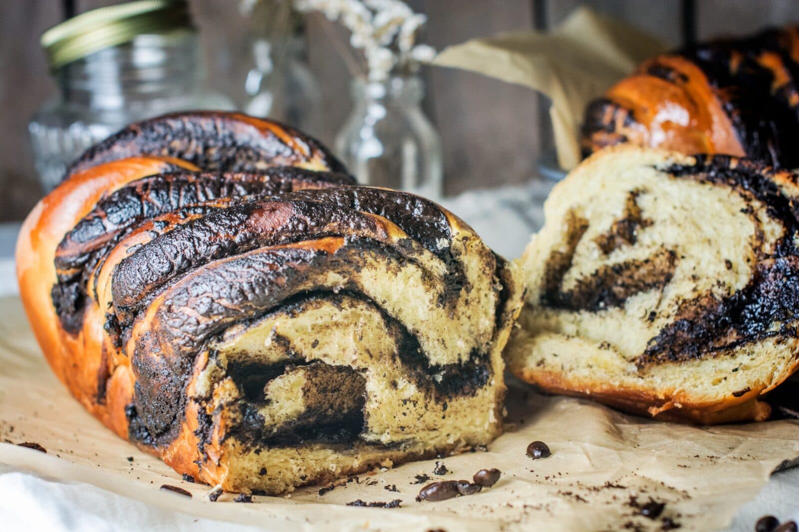 A close-up of sliced coffee babka on parchment paper, showing swirls of chocolate and coffee filling in the soft, golden-brown bread. A jar and glass vases are blurred in the background.