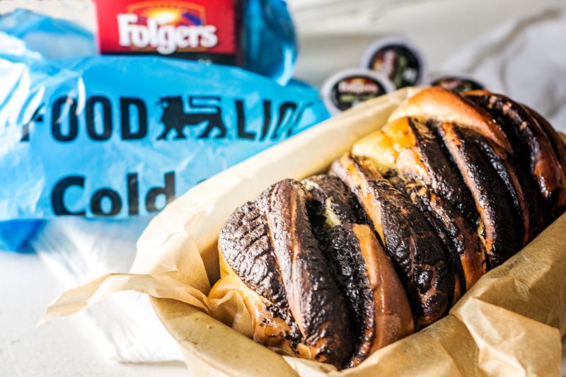 A loaf of Coffee Babka bread in parchment paper sits in front of a blue Good & Gather cold cup package and a container of Folgers coffee, with coffee pods in the background.
