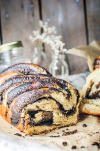 A close-up of a sliced loaf of coffee babka bread on parchment paper, with rich swirls of chocolate filling visible. In the background, glass jars and dried flowers rest on a rustic wooden surface.