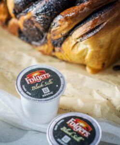 Three Folgers Black Silk Keurig coffee pods rest on a surface near scattered coffee beans and a freshly baked Coffee Babka loaf on parchment paper. The focus is on the rich coffee pods and the beautifully braided bread.