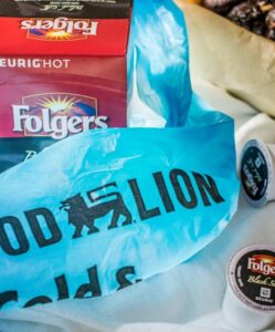 A Food Lion plastic bag covers a box of Folgers Black Silk Keurig coffee pods, with several pods scattered nearby and a coffee babka in a paper tray in the background.