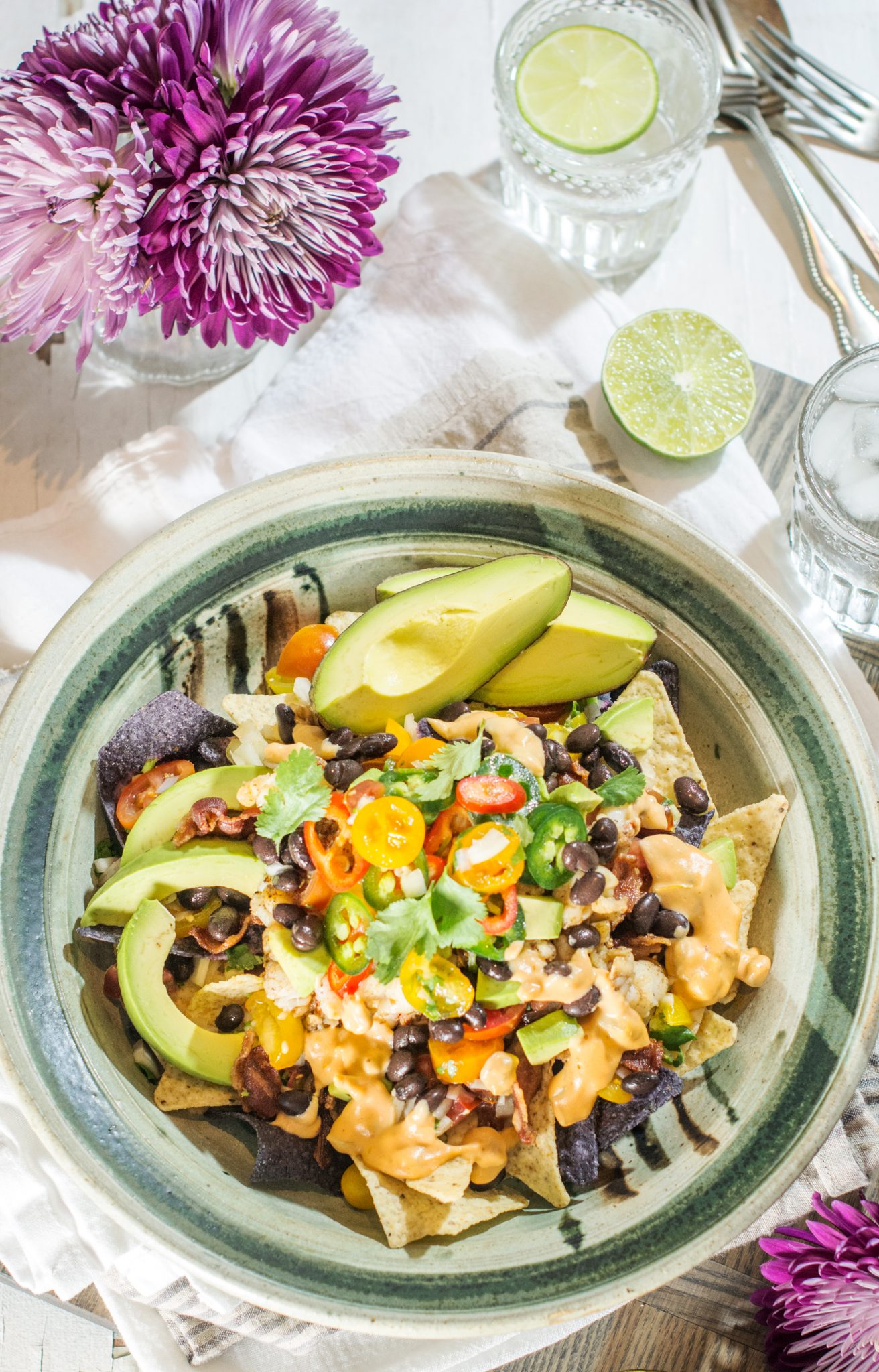 A plate of Lobster Nachos topped with black beans, cheese sauce, sliced avocado, cherry tomatoes, jalapeños, and cilantro. The dish is served on a rustic plate with lime halves, flowers, and glasses of water nearby.