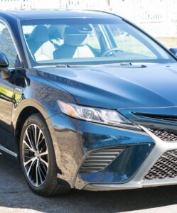 A shiny blue Toyota Camry sedan is parked on a driveway in daylight, with a white fence and grass in the background—perfect for enjoying Lobster Nachos nearby.