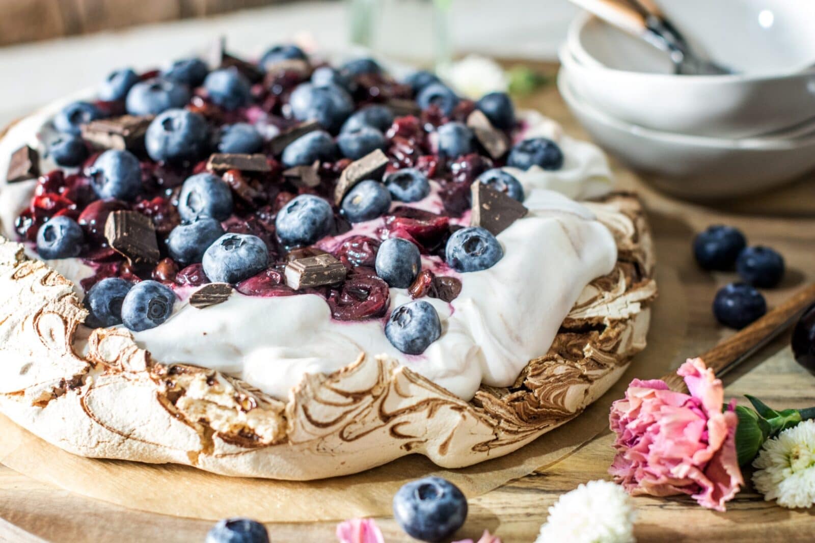 A Chocolate Pavlova dessert topped with whipped cream, blueberries, chopped chocolate, and berry sauce sits on a wooden board, surrounded by fresh flowers and stacked white bowls with spoons in the background.
