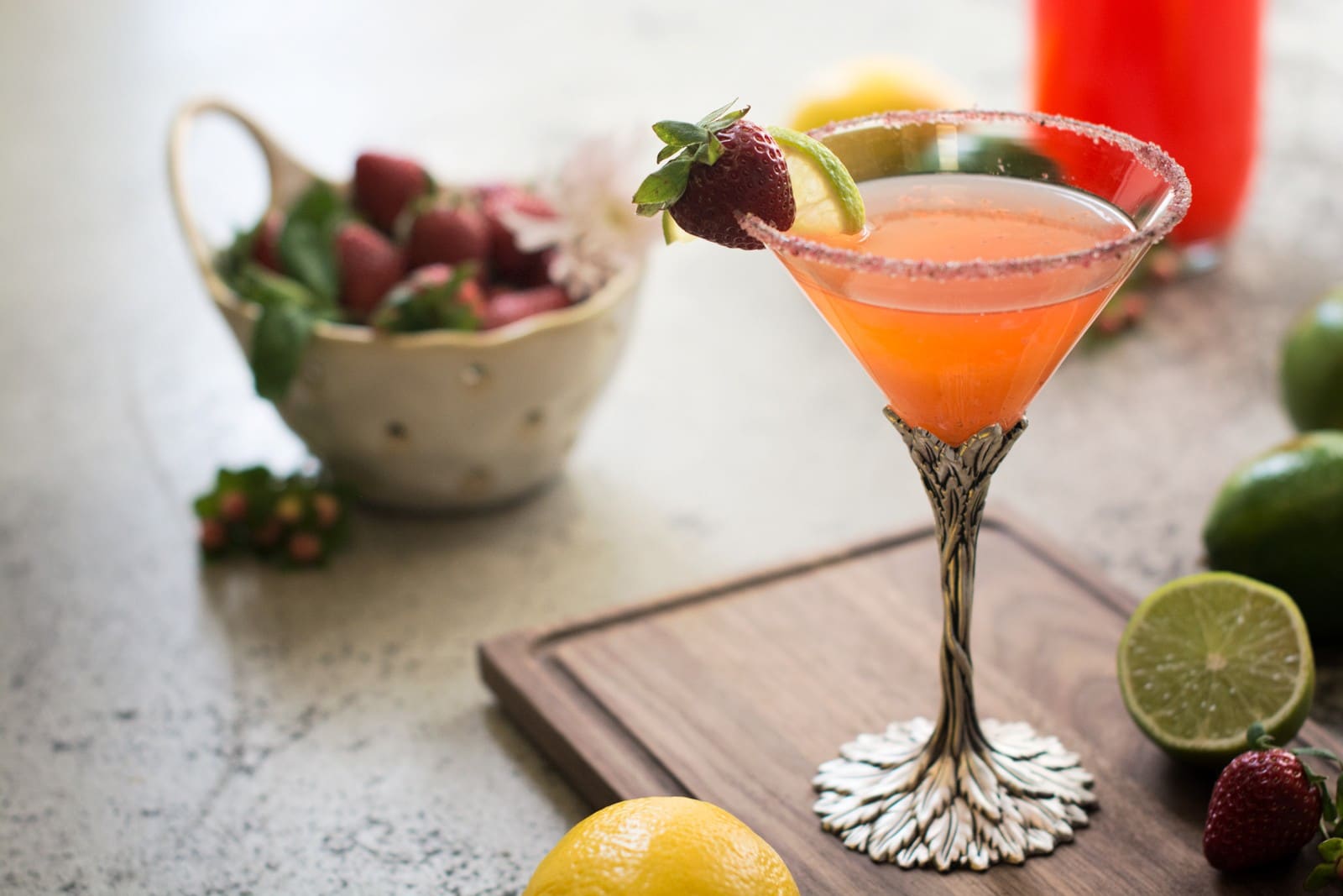 A Strawberry Daiquiri in an ornate glass, garnished with a strawberry and lime slice, sits on a wooden board surrounded by fresh strawberries, lemons, and limes.